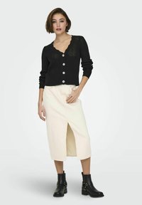 Black knitted cardigan with a scalloped neckline, button closure, and long sleeves, paired with a cream midi skirt featuring a front slit. Black ankle boots.
