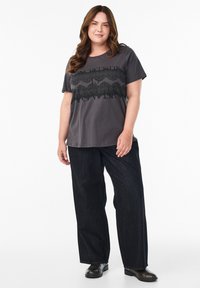 Grau T-Shirt mit einem strukturierten schwarzen Spitzenmotiv über der Brust, kombiniert mit lockeren dunklen Jeans und schwarzen Stiefeletten.