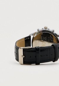 Uhr mit schwarzem Lederarmband und Metallschließe, Rückseite eines silbernen Gehäuses aus Edelstahl auf weißem Hintergrund sichtbar.