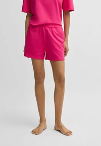 Rosa bomullsshortsen med elastisk midja, med en enkel design och mjuk textur. Modellen står barfota mot en neutral bakgrund.