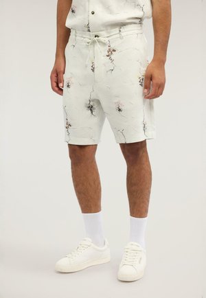 Mann trägt weiße Shorts mit Blumenmuster, weiße Socken und weiße Turnschuhe, steht vor einem schlichten hellen Hintergrund.