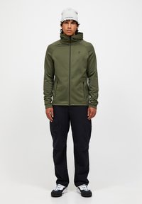 Giacca in pile verde con zip frontale, colletto alto e due tasche laterali, abbinata a pantaloni cargo neri e sneakers bianche.