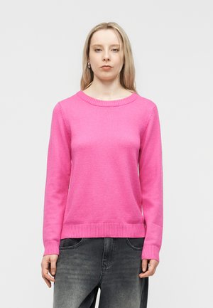 Femme portant un pull à manches longues rose vif et un jean gris foncé, debout devant un fond gris clair uni.