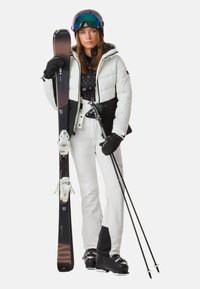 Vrouw in een witte en zwarte skikleding, met ski's en stokken in de hand, die een helm, goggles en zwarte skischoenen draagt, tegen een witte achtergrond.