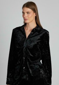 Camisa de terciopelo negro con mangas largas, botones en la parte delantera y cuello redondeado. Presenta una sutil textura y acentos de botones brillantes.
