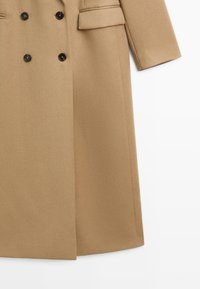 Double-breasted camel coat made of textured fabric. Features black buttons, slim lapels, and a straight-cut hem. Tailored pockets included.