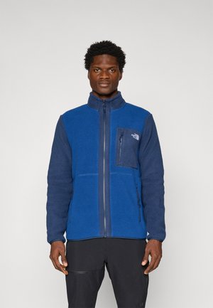 Blue fleece jacket with a high collar, zip closure, and a chest pocket. Features a mix of textures and colors in a soft, warm material.
