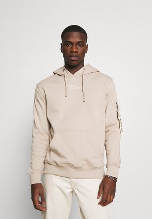 Beige Kapuzensweatshirt mit einer Fronttasche, Kordeln und einem Stoffaufnäher am linken Ärmel, aus weichem Baumwollmaterial gefertigt.