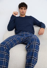 Cămașă cu mânecă lungă de culoare bleumarin, asortată cu pantaloni de pijama cu carouri albastre și accente albe, așezată pe o canapea texturată, de culoare deschisă.
