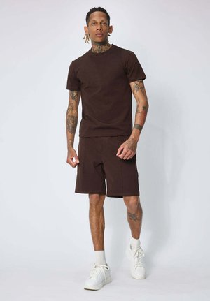 Homme avec des tatouages portant un t-shirt marron foncé et un short, des chaussettes blanches et des baskets blanches, debout contre un fond clair uni.