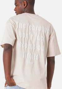 Beige bomulds T-shirt med korte ærmer, prydet med hævet hvid tekst på ryggen: "De frygter vi ikke konfronterer bliver vores grænser."