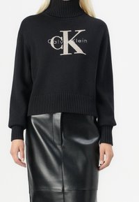 Pull noir en tricot avec col roulé, poignets et ourlet côtelés, présentant un logo "CK" beige contrastant et le texte "Calvin Klein" sur le devant.