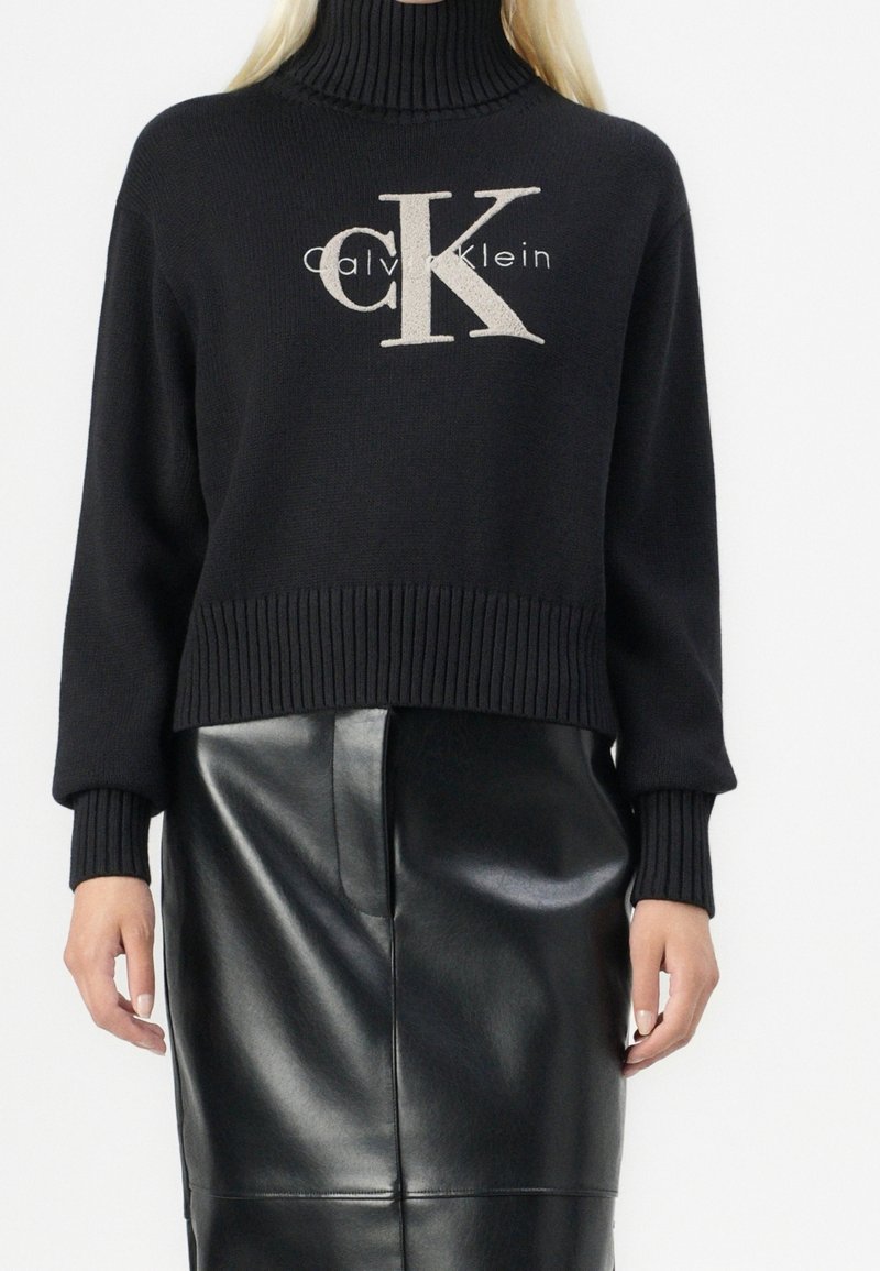 Pull noir en tricot avec col roulé, poignets et ourlet côtelés, présentant un logo "CK" beige contrastant et le texte "Calvin Klein" sur le devant.