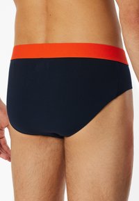 Herrenunterhosen aus navyblauem Stoff mit einem orangen elastischen Bund, die eine enge Passform und eine glatte Textur bieten. Keine sichtbaren Muster oder Logos.