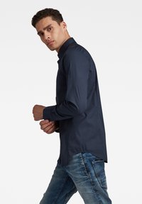 Camicia blu navy con bottoni, vestibilità slim, maniche lunghe e orlo curvo, abbinata a jeans chiari strappati.