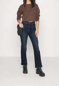 Mujer vestida con un suéter corto marrón, jeans de tiro alto azul oscuro, botas de plataforma negras hasta el tobillo y sosteniendo un bolso bandolera texturizado negro.
