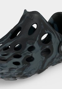 Μαύρο παπούτσι χωρίς κορδόνια με πολλαπλές άνισες οπές αερισμού και υφή στην επιφάνεια, με στρογγυλεμένη μύτη και ρυθμιζόμενο λουράκι στη φτέρνα.