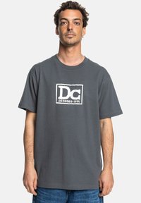 Graues Baumwoll-T-Shirt mit kurzen Ärmeln, das ein weißes rechteckiges Logo in der Mitte aufweist. Einfaches, lässiges Design.