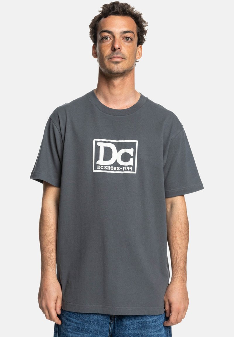Graues Baumwoll-T-Shirt mit kurzen Ärmeln, das ein weißes rechteckiges Logo in der Mitte aufweist. Einfaches, lässiges Design.