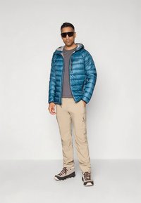 Tirkīza krāsas pūkaina jaka ar pelēku kapuci, valkāta pār pelēku T-kreklu. Bēšas krāsas darbagabalu bikses un brūnas pārgājienu zābaki, stāvot pret baltu fonu.