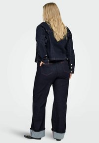 Temno modra denim jakna in razširjene kavbojke z lahkimi manšetami iz denima. Ima rdeče šive in kovinske gumbe. Pogled od zadaj.