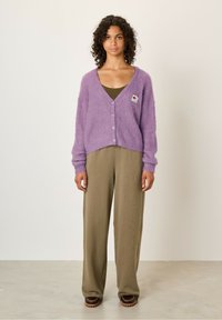 Lila fuzzy cardigan met knopen, aangevuld met een groene top eronder. Wijdvallende kaki broek en bruine schoenen maken de outfit compleet.