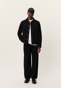 Μαύρο τζιν μπουφάν με κουμπιά, παντελόνι με φαρδύ πόδι και μαύρο καπέλο με κίτρινο κείμενο. Ομαλή υφή, μίνιμαλ σχεδιασμός.