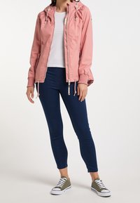 Lichtroze zip-up jack met capuchon, trekkoorddetails, geribbelde manchetten, gedragen over een witte top en gecombineerd met blauwe skinny jeans en groene sneakers.