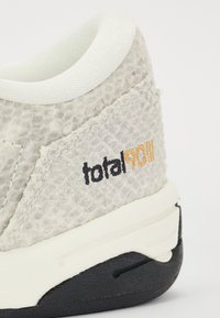 Zapatilla gris claro con parte superior texturizada, cuello blanco y suela negra. Presenta un logo bordado "total90" en negro y oro.