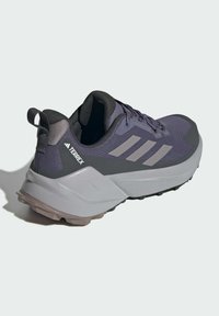 Chaussures de plein air grises avec un upper texturé et respirant ; talon renforcé ; et logo à trois rayures bien visible sur le côté.