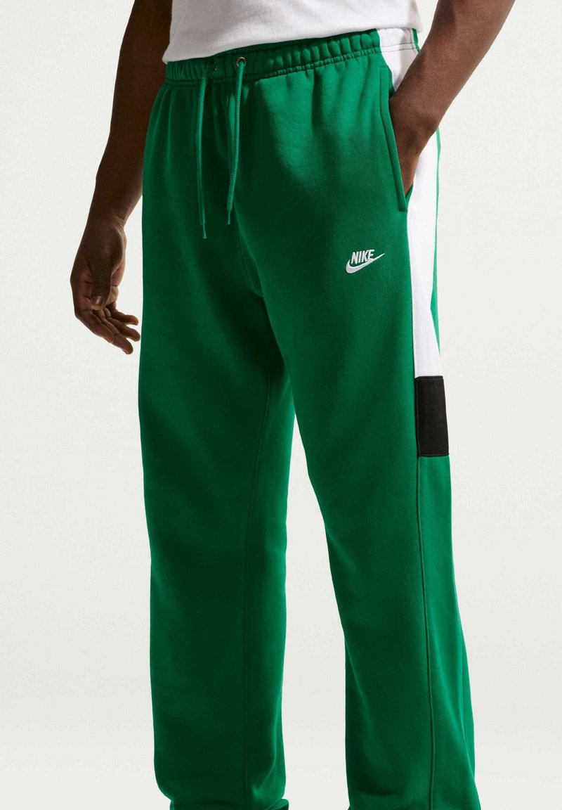 Uomo che indossa pantaloni della tuta Nike verdi con strisce laterali bianche e nere, una mano in tasca, camicia bianca parzialmente visibile.
