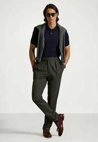 Mężczyzna w okularach przeciwsłonecznych, granatowej koszulce polo, szarym swetrze zarzuconym na ramionach, szarych spódnicach w kant i brązowych skórzanych butach, stojący z rękami w kieszeniach.