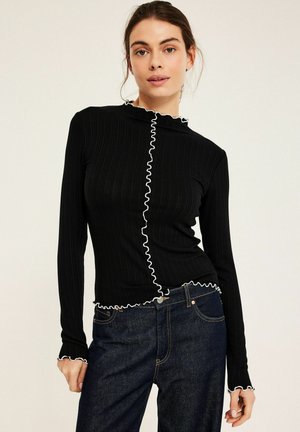 Frau trägt ein eng anliegendes schwarzes Langarmshirt mit weißem Rüschenbesatz und dunkelblaue Jeans vor einem einfarbigen hellen Hintergrund.