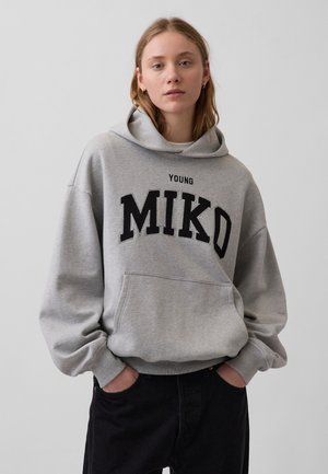 Ung kvinde iført en grå hoodie med teksten "YOUNG MIKO", hænderne i lommerne, står op ad en ensartet lysegrå baggrund.