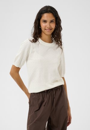 Femme aux cheveux bouclés foncés sourit, portant un haut en maille blanc à manches courtes et un pantalon marron ample avec cordon, debout devant un fond uni.