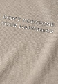 Tela beige con una superficie texturizada que presenta letras en relieve plateadas que dicen: "NO POSTERGUES TU FELICIDAD."