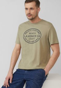 Oliwkowa bawełniana koszulka z krótkim rękawem, z okrągłym czarnym nadrukiem graficznym z tekstem. Połączona z niebieskimi dżinsami.