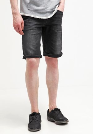 Homme portant un short en denim noir retroussé, un t-shirt gris et des baskets noires, debout avec une main dans la poche sur fond blanc.