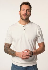 Camicia bianca a maniche corte con scollo rotondo e patta con tre bottoni. Presenta un orlo a costine e una trama semplice, abbinata a jeans di denim.