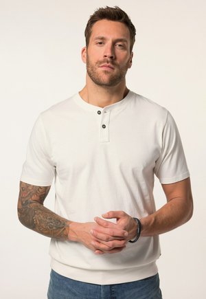 Camisa blanca de manga corta con cuello redondo y tapeta de tres botones. Presenta un dobladillo acanalado y textura lisa, combinada con pantalones vaqueros.