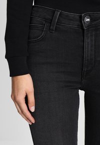 Pantalones vaqueros de denim negro con un ajuste slim, diseño estándar de cinco bolsillos y cierre de botón de marca. Textura sutil con un ligero estiramiento.