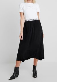 Gonna nera midi con fascia elastica e logo di marca. Abbinata a una t-shirt bianca aderente e stivaletti neri alla caviglia con punte metalliche.