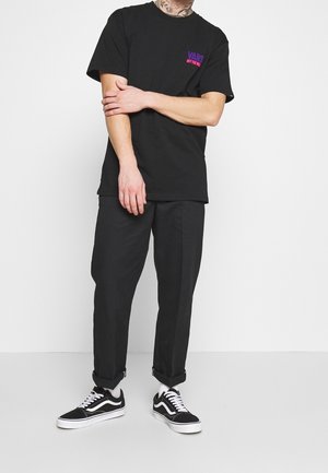 Persoon die een zwart Vans T-shirt met paars-roze logo draagt, zwarte omgeslagen broek, witte sokken en zwarte Vans sneakers, staand tegen een witte achtergrond.