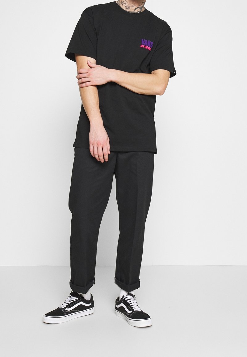 Personne portant un t-shirt Vans noir avec un logo violet et rose, un pantalon noir retroussé, des chaussettes blanches et des baskets Vans noires, debout devant un fond blanc.