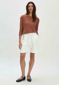 Gestreiftes braunes Langarmoberteil mit einem pinken Kragen, kombiniert mit weißen Shorts, die zwei vordere Taschen und einen Kordelzugbund haben. Schwarze Ballerinas.