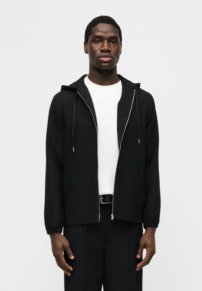 Νέος άνδρας που φοράει μαύρο φούτερ με φερμουάρ πάνω από λευκό πουκάμισο και μαύρο παντελόνι, στέκεται μπροστά σε απλό λευκό φόντο.