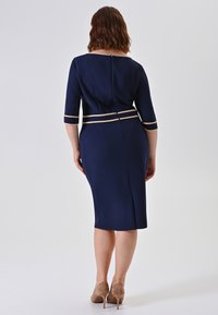 Frau mit welligem braunem Haar, die ein marineblaues knielanges Kleid mit beigem Besatz trägt, mit dem Rücken zur Kamera stehend und beigen High Heels.