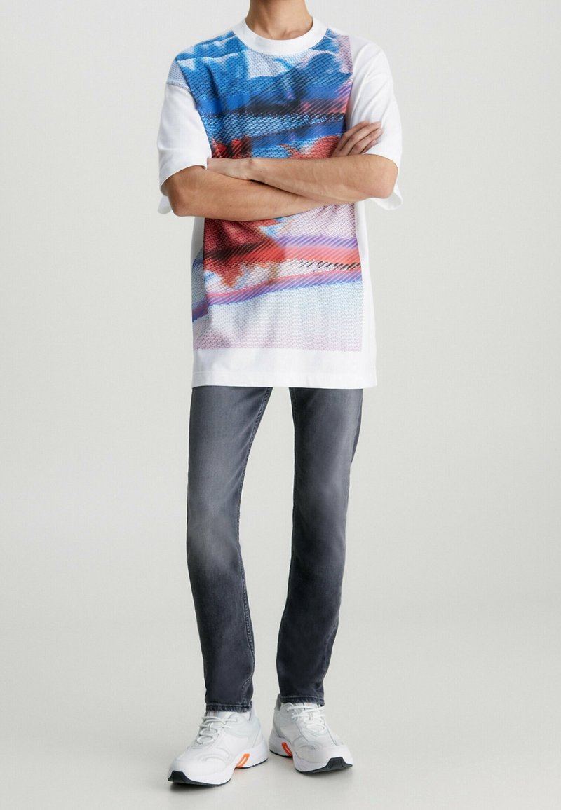 Kortärmad vit t-shirt med ett blått och rött grafiskt mönster, kombinerad med grå jeans och vita sneakers med orangea detaljer.