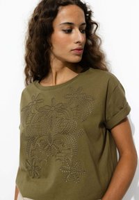 T-shirt en coton vert olive avec des manches retroussées, ornée de motifs floraux en relief sur le devant. Texture douce, coupe décontractée.