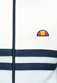 Λευκό τζάκετ με φερμουάρ και ναυτικές ρίγες, με πορτοκαλί και κίτρινο κυκλικό λογότυπο. Κατασκευασμένο από υφασμα με υφή, διαθέτει κεντρικό φερμουάρ και διακοσμητικές ραφές.